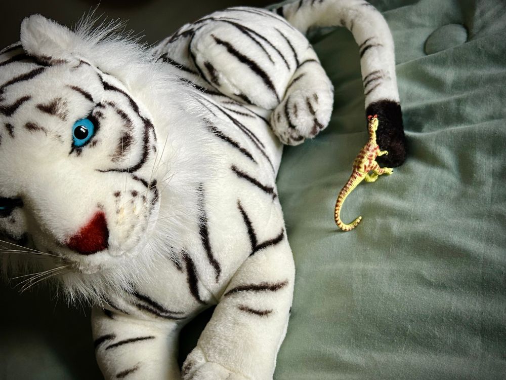 A green Dilophosaurus with brown spots holds a giant Siberian tiger (white with black stripes) as he lounges on a green winter puff atop a bed. The tiger has bright blue eyes and a red nose.