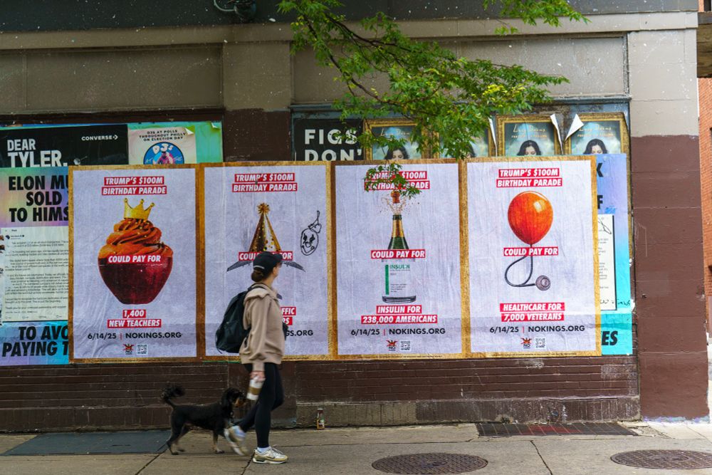 A person walks a black dog past a row of four large posters on a brick wall. Each poster critiques the $100 million cost of Trump’s birthday parade by highlighting alternative uses for the money. The posters read:
	1.	“Trump’s $100M Birthday Parade could pay for 1,400 new teachers” with an image of a frosted cupcake with a crown.
	2.	“Trump’s $100M Birthday Parade could pay for 700 air traffic controllers” with a party hat and airplane illustration.
	3.	“Trump’s $100M Birthday Parade could pay for insulin for 238,000 Americans” with an insulin vial styled as a champagne bottle.
	4.	“Trump’s $100M Birthday Parade could pay for healthcare for 7,000 veterans” with a red balloon and stethoscope.
The date “6/14/25” and the URL “NOKINGS.ORG” appear at the bottom of each poster.