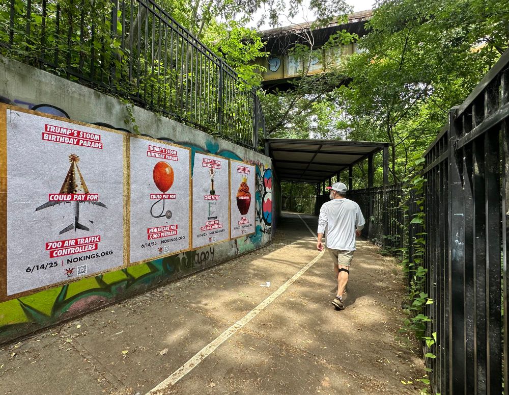 A man in a white shirt and cap walks up a graffiti-lined pedestrian path. On the cement wall to his left are four large posters criticizing Trump’s $100M birthday parade:
	1.	A party hat shaped like a plane: “Could pay for 700 air traffic controllers.”
	2.	A balloon and stethoscope: “Could pay for healthcare for 7,000 veterans.”
	3.	A champagne-bottle insulin vial: “Could pay for insulin for 238,000 Americans.”
	4.	A cupcake with a crown: “Could pay for 1,400 new teachers.”
The date and website “6/14/25 | NOKINGS.ORG” are on each.
