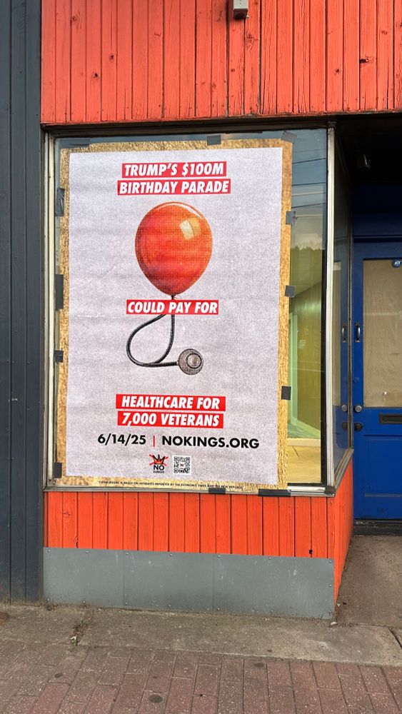 A single large poster is taped to the window of a boarded-up building with red and black siding. It reads: “Trump’s $100M Birthday Parade could pay for healthcare for 7,000 veterans,” with a red balloon and stethoscope graphic. Below the main text, the date “6/14/25” and URL “NOKINGS.ORG” are printed above the “No Kings” logo and QR code.