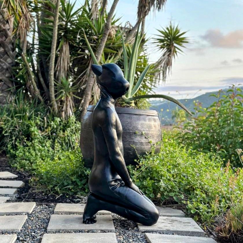 Fern sitting on his heels among plants and a large pot. He's wearing a smooth gray silicone suit and a matching pup hood with only a mouth hole
