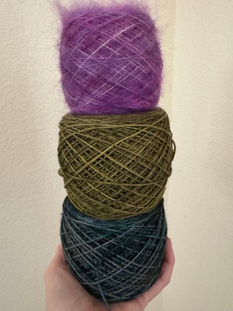 Three cakes of yarn held in someone’s left hand. The top cake is purple and is stacked on top of a moody green cake, which is stacked on top of a grey blue cake of yarn.