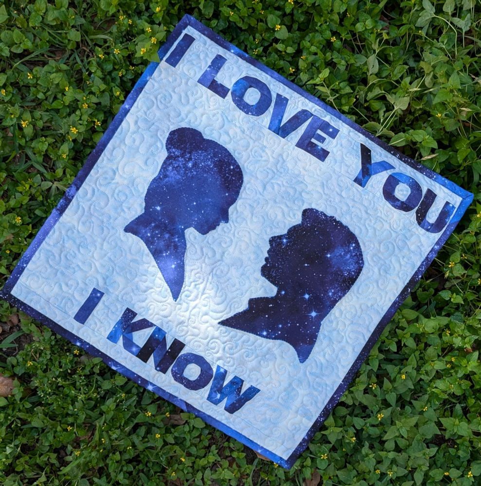 Small Star Wars themed quilt with a light blue background. Has the words "I love you, I know" in dark blue celestial fabric framing the top and bottom. Silhouettes of Princess Leia and Han Solo facing each other in the center. 
