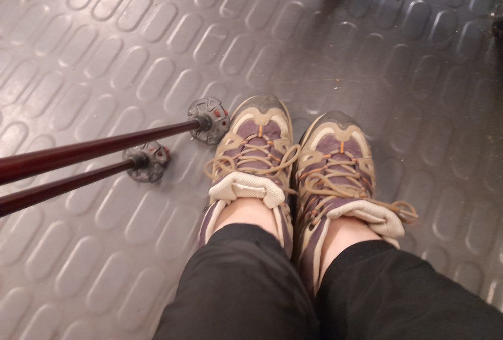 Photo orientée vers le sol de pieds chaussés de chaussures de randonnée et des bâtons de randonnée dépliés, prise dans le RER.