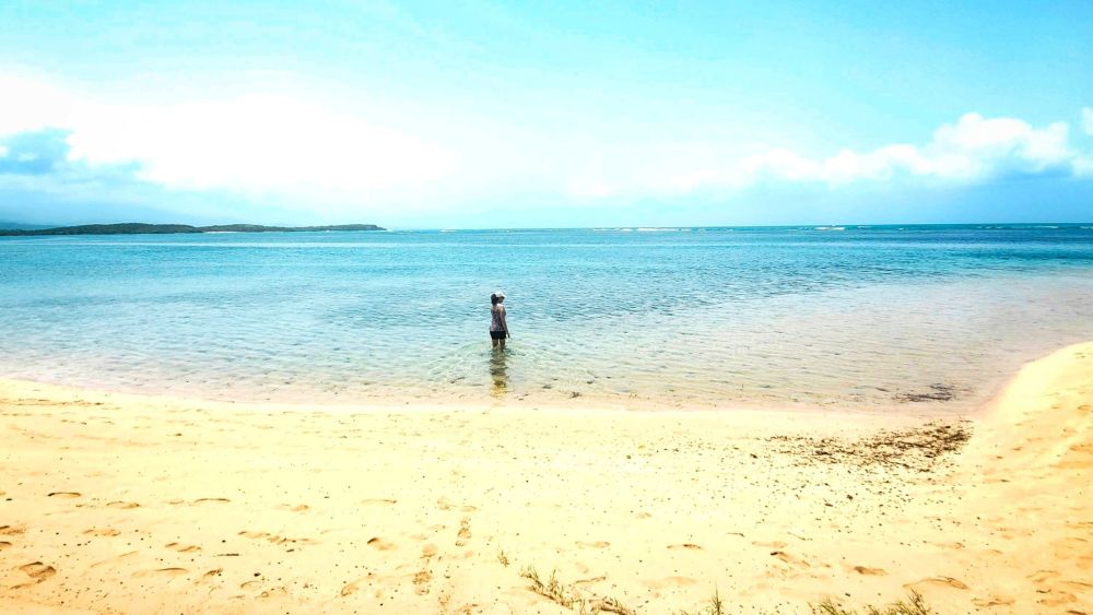 I'm standing in shallow ocean water under a light blue sky, gazing at the horizon with golden sand in the foreground and distant islands in the background.
