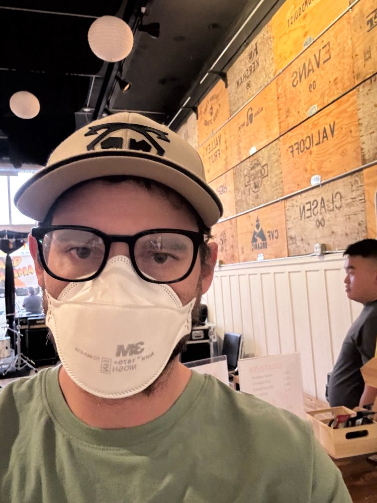 Selfie of me(a white male) in a 3M respirator, glasses, and a hat. In the background is an event space and a couple of humans. 