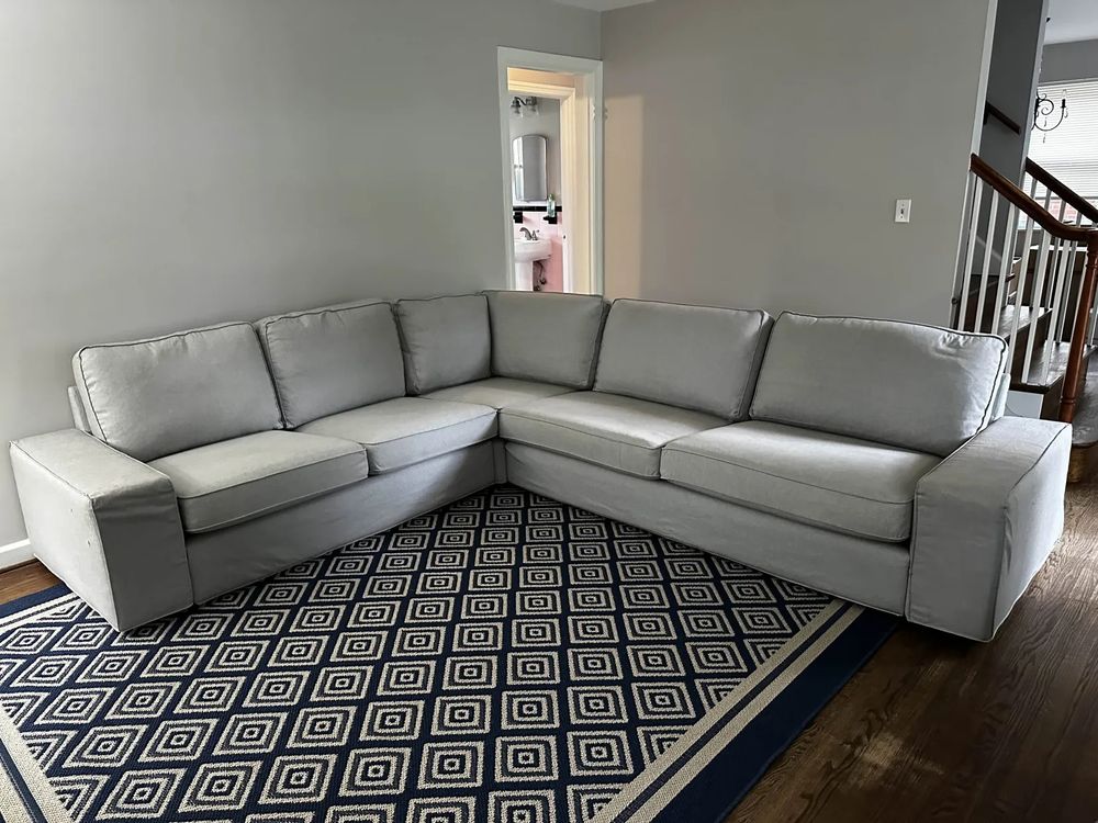 image is of a sectional sofa in gray; it is partially covering a navy and tan rug that has a symmetrical diamond pattern. The room is painted a neutral gray and visible in the background is a doorway to a hallway and a stairway railing 