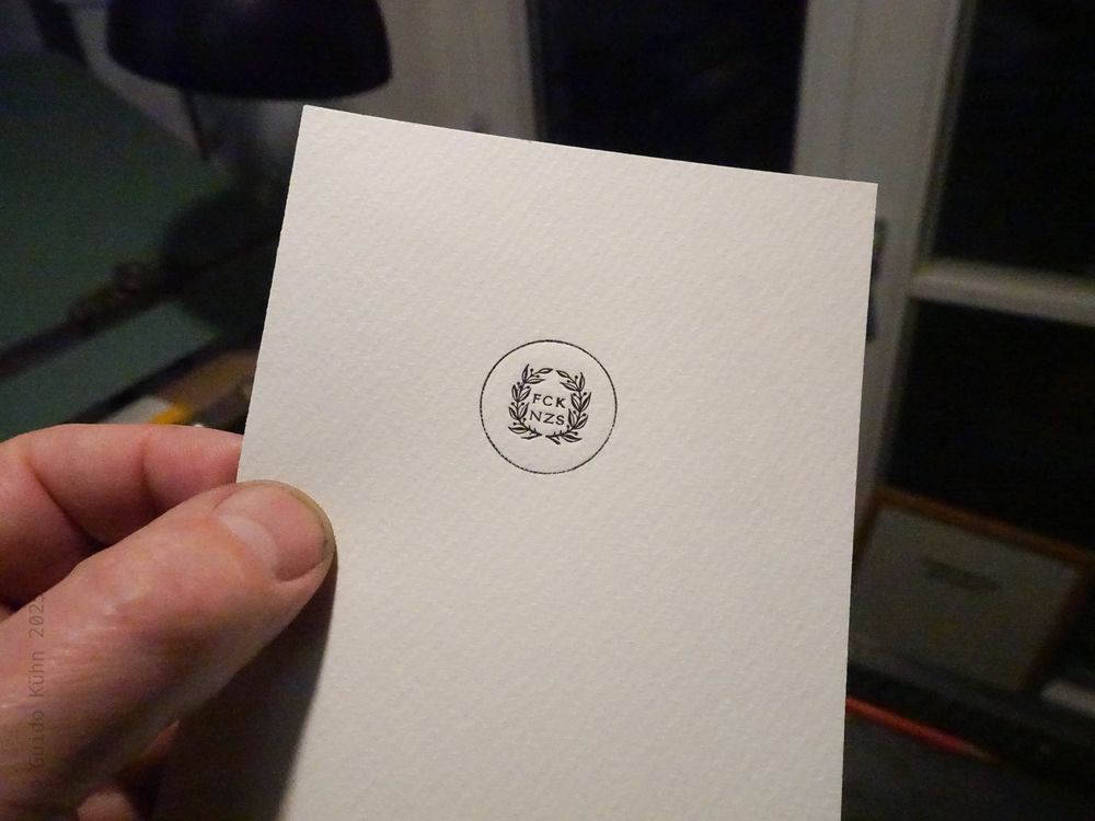 Foto des Abzuges einer Bleisatzform, bei der ein paar Loorbeeräste um zwei Zeilen mit den sechs Buchstaben FCK NZS gesetzt und durch einen umlaufenden Kreis gefasst werden. 