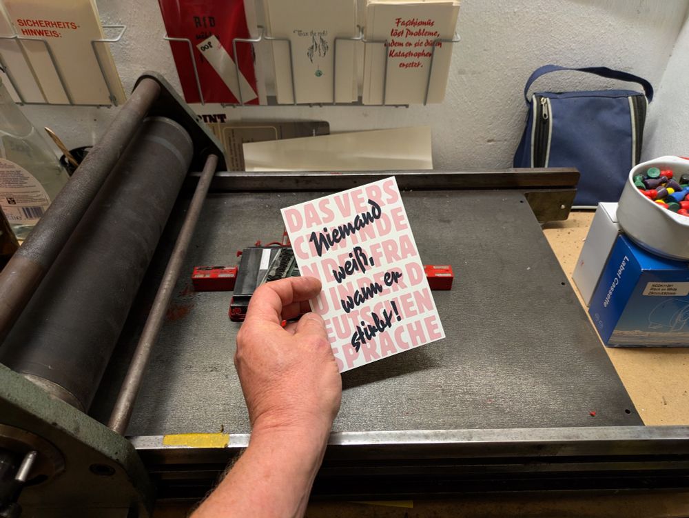 Foto meiner Hand einen kartengroßen, zweifarbigen Abzug in der Hand haltend. Blassurot und formatfüllend steht im Hintergrund kompress durchlaufend auf Block gesetzt: Das verschwinden der Frau in der Deutschen Sprache. Davor in schwarzer, diagonal über vier Zeilen gesetzter Pinselschrift die Aussage: Nimand weiß, wann er stirbt!

Die Schriftarten sind rot: Block und schwarz: Reporter