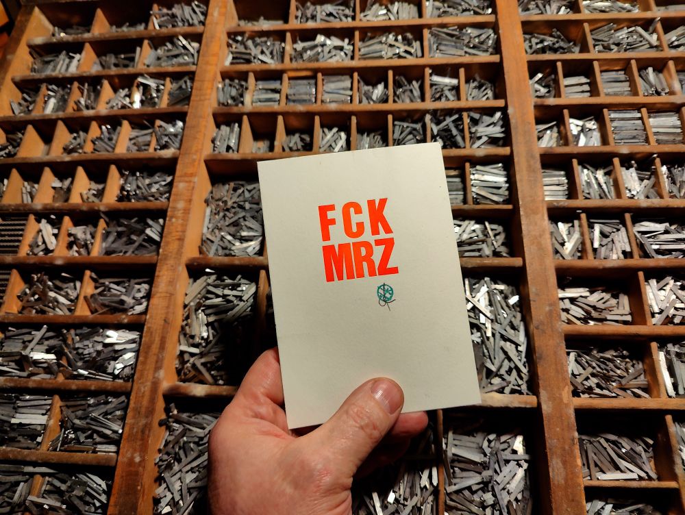 Foto_Hand hält Karte in Bleisatz vor Setzkasten. Auf der Karte steht in Leuchtorange FCK MRZ