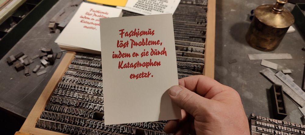 Foto meiner Hand die eine in Bleisatz gesetzte und rot gedruckte Karte vor den Setzkasten hält. Der Text lautet: Faschismus löst Probleme, indem er sie durch Katastrophen ersetzt. Die Schrift ist die Pinselschrift Reporter, eine beliebte Werbe- und Plakatschrift der 1930er und 40er.