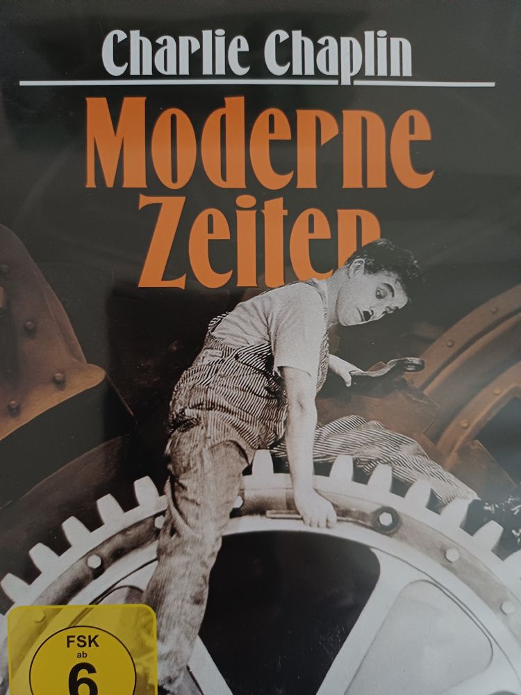 DVD 'Moderne Zeiten' von u. mit Charlie Chaplin.  Chaplin sitzt auf einem großen Zahnrad. Im Hintergrund Maschine, in die er eingeklemmt ist.
