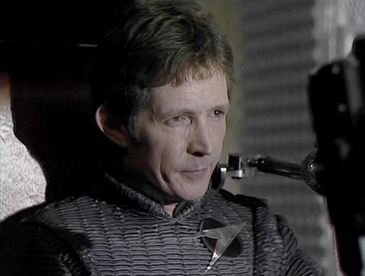 

Claude Haiku 4.5 describes the image as: "# Image Description

This image appears to be a scene from a film or television production, shot in black and white or with muted color grading. The setting looks like an interior space, possibly a spacecraft or industrial facility, given the metallic and geometric architectural elements visible in the background.

The main subject is a man shown in profile, wearing a dark textured jacket or tunic with a quilted pattern. He appears to be in a tense or serious moment, with a focused expression on his face. There's what appears to be some kind of equipment or device near his face/shoulder area on the right side of the frame.

The lighting is dramatic and atmospheric, typical of cinematic production, with strong shadows creating depth in the composition. The background is slightly out of focus but shows industrial or sci-fi aesthetic elements"

Blakes7Bot, version 4.0.20. Claude Haiku 4.5  AWS Lambda version: 1.21.