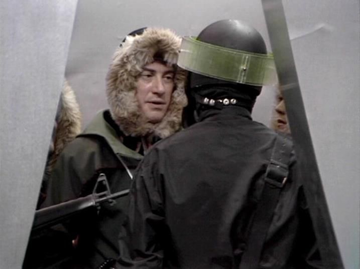 

Claude 3.7 describes the image as: "This image appears to be from a science fiction television series from the late 1970s or early 1980s. It shows a tense confrontation scene in what looks like a sterile, institutional corridor or doorway. 

On the left is a person wearing a fur-trimmed parka hood, facing off against what appears to be a security guard or soldier in dark clothing wearing a helmet with a visor. The guard is holding what looks like a weapon.

The scene has the distinctive visual style of British sci-fi productions from that era, with minimalist set design and a somewhat muted color palette. The lighting and film quality have that characteristic vintage television production look, suggesting this is from a classic science fiction series.

The confrontation appears to be a dramatic moment, possibly a standoff between a rebel or civilian and authoritarian forces."

Blakes7Bot, version 3.9.31. AWS Lambda version: 1.14.