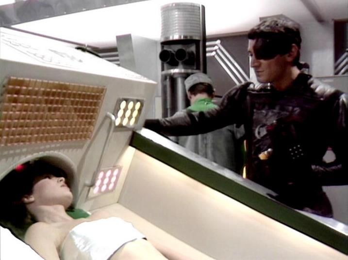 

Claude 3.7 describes the image as: "This image appears to be from a science fiction television series, showing a futuristic medical or examination scene. In the foreground, there's a person lying on what looks like a medical bed or examination table, wearing what appears to be a white garment. Standing nearby is another individual dressed in dark clothing with some sort of eye covering or visor, reaching toward a control panel with illuminated sections. The setting has a distinctly retrofuturistic aesthetic with metallic surfaces, technical equipment, and distinctive lighting panels. The scene appears to be taking place in what might be a spaceship or advanced facility, with industrial-looking design elements typical of science fiction productions from that era."

Blakes7Bot, version 3.9.31. AWS Lambda version: 1.14.