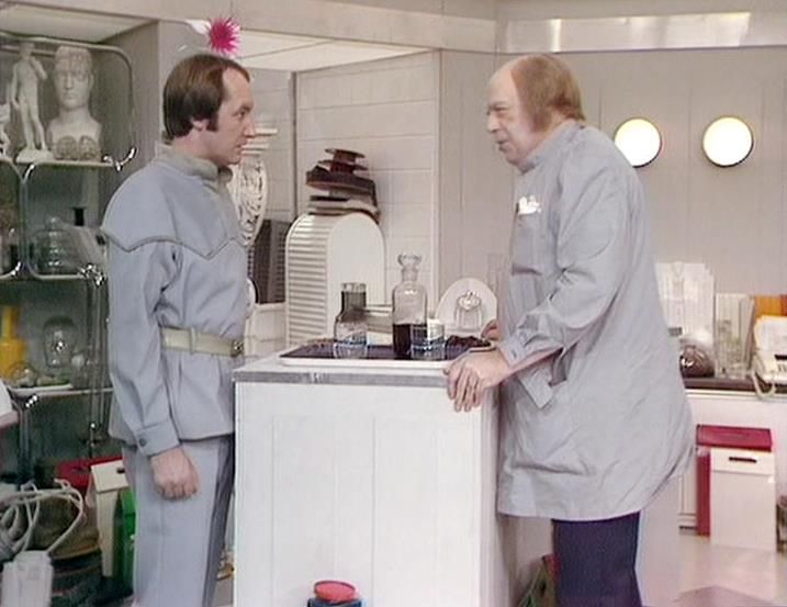 

Claude 3.7 describes the image as: "This image shows a scene set in what appears to be a futuristic medical or laboratory facility with a clean, white interior design. Two people in gray uniform-like clothing are engaged in conversation across a white counter or examination table. One person on the left has a distinctive pink decoration or antenna on their head, while the person on the right has a more authoritative stance.

The clinical setting includes glass shelving with medical equipment, bottles of liquids on the counter between them, and circular lighting fixtures visible on the wall. The stark, minimalist aesthetic is typical of science fiction productions from this era, with the costumes suggesting some kind of institutional or organized setting.

The scene appears to capture a moment of serious discussion in what might be a medical bay or scientific facility."

Blakes7Bot, version 3.9.31. AWS Lambda version: 1.14.
