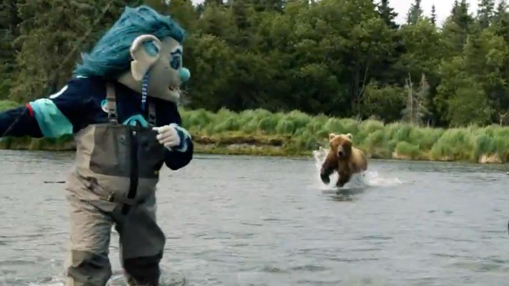 The mascot for the NHL team the Seattle Kraken running from a wild grizzly bear that is chasing it across a body of shallow water during a photo shoot in Alaska.