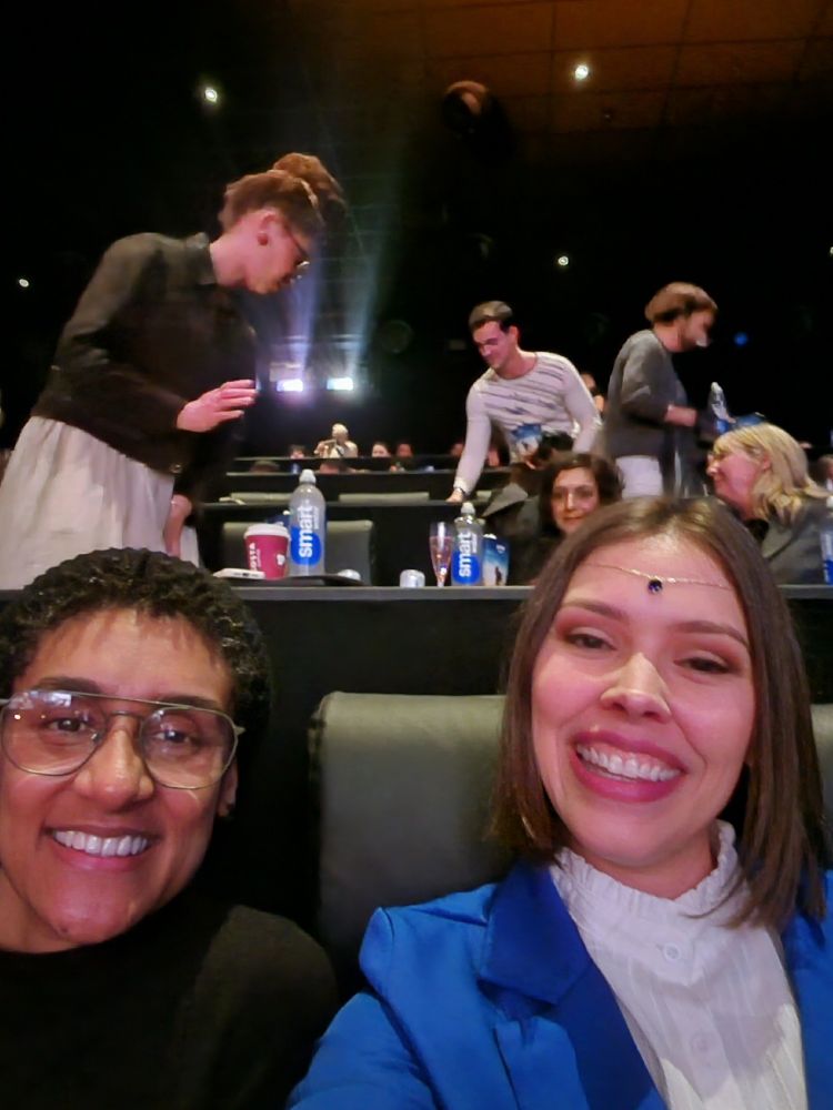 Selfie of two women smiling at the camera and sitting in leather theater chairs. In the row behind them, Meera Syal is sitting and Ayoola Smart is getting to their seat. 