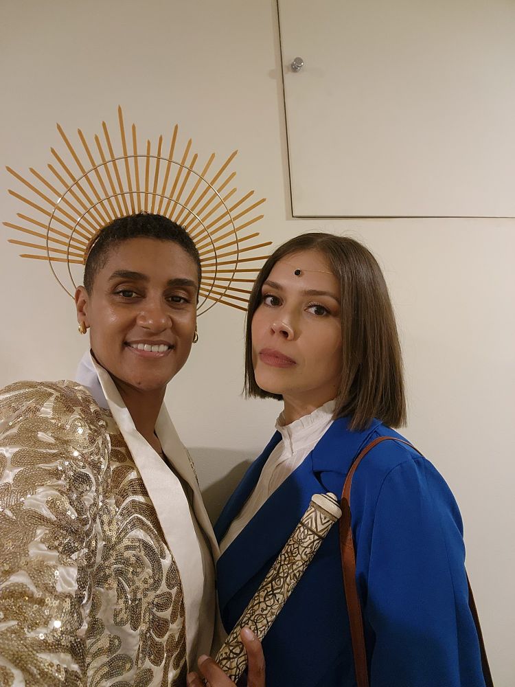 Two women pose in cosplay outfits of Siuan and Moiraine. The one on the left wears a gold blazer and halo spikey gold crown, and holds an oath rod. The one of the right wears a blue blazer with white shirt under and has a blue jewel on her forehead.
