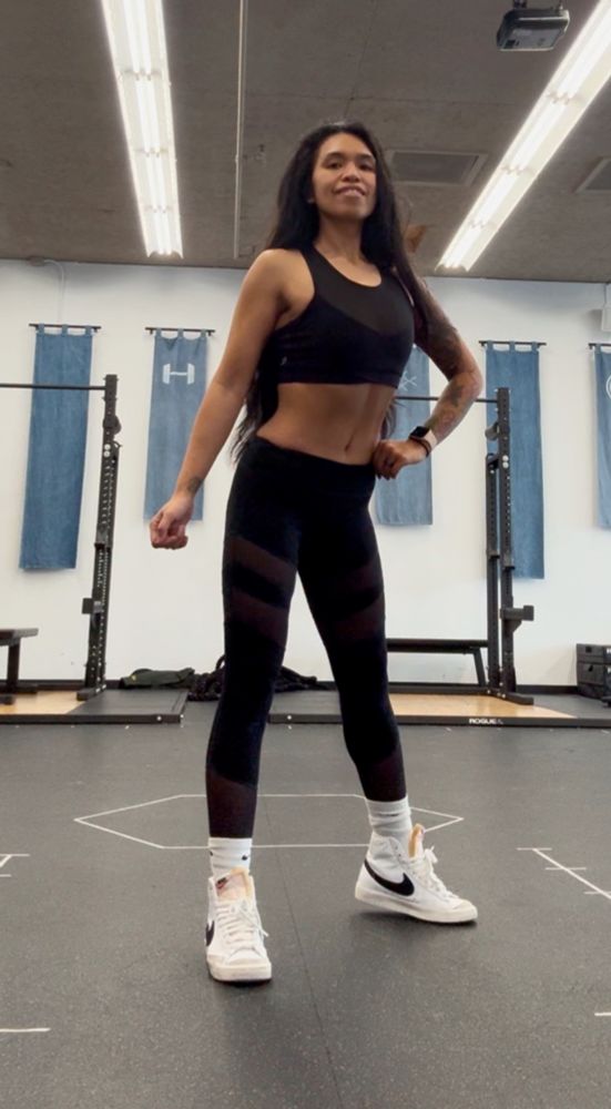 A somewhat athletic-looking woman in  the middle of a gym posing with her left hand on her hip and right hand straight out to the side. Her feet are slightly more than hip width apart.