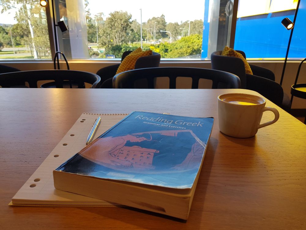 JACT Reading Greek textbook sitting on a blank writing pad with a metal pen crossed on it. There is a coffee beside the book.
In front of the table, large windows open onto the massive yellow lettering of an Ikea store mounted on a blue background. In the middle distance, there are trees with roads crossing between them. Bliss.