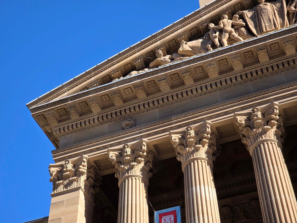 Three fluted Corinthian columns on the Brisbane City Hall, with an acute triangle of the neo-classical pediment visible above. The sculptures on the pediment were carved by Daphne Mayo in 1027-30, featuring images of white settlement 
The plain stone column on the left side, an extension of the end wall, also features a Corinthian capital.