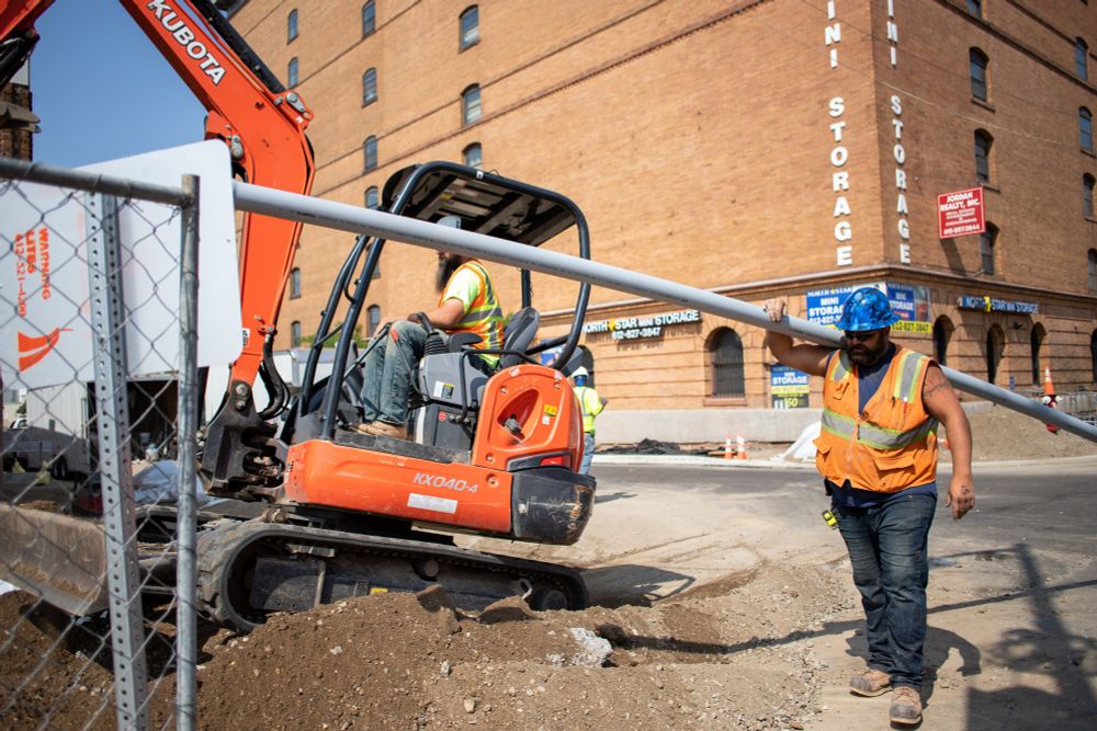 A construction worker carries a pipe while another worker operates a backhoe in the background