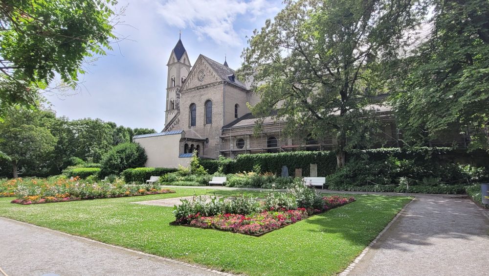 Blick in die Gartenanlage rechts neben der Basilika St. Kastor Koblenz. Vorne eine akkurat gepflegte rechteckige Rasenfläche, in die Blumenbeete mit bunten Blumen eingelassen sind. Im Hintergrund die Seitenansicht der Basilika.
