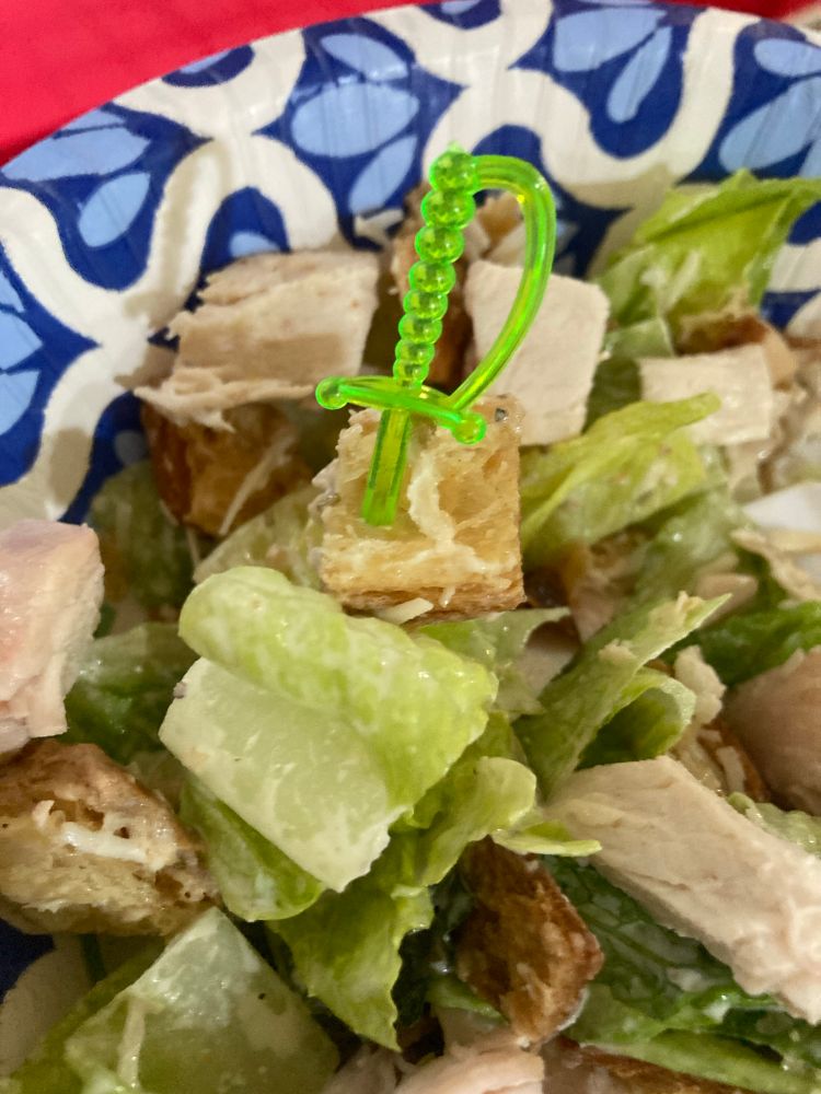 This photo shows a bite portion of Caesar salad impaled by a green plastic sword toothpick held above a blue and white paper bowl of the salad on a red tablecloth