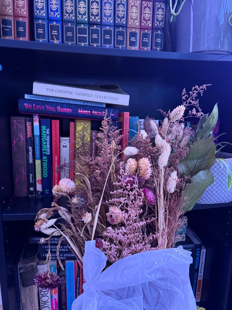 background: a snippet of Faye’s (book) library 
foreground: a dried flower bouquet wrapped in a white garbage bag 