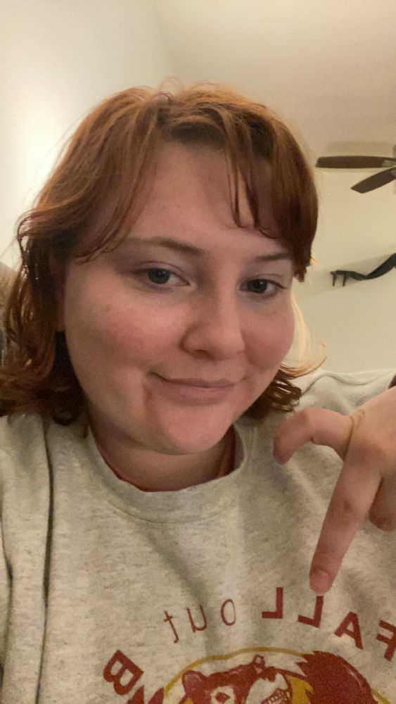 me, a white person with red hair closed mouth smiling while making half a heart with their fingers. i have on an off white fall out boy sweater