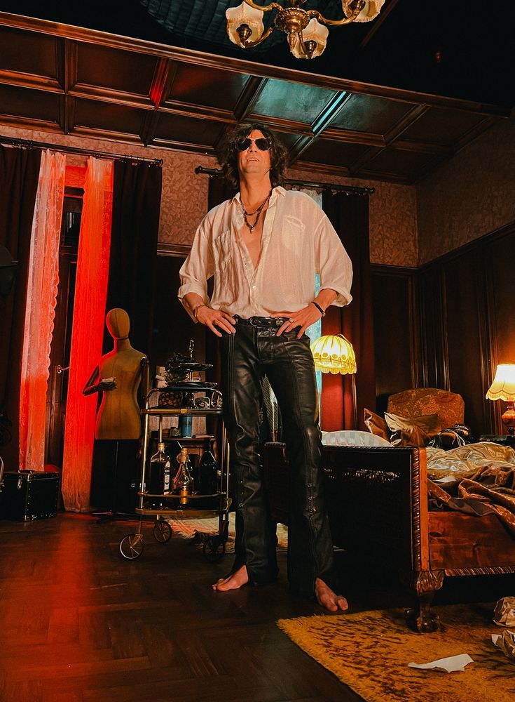 Ilkka Villi as Thomas Zane in Room 665. He's dressed in a romantic white shirt with a lot of the buttons undone, and long leather trousers. His feet are bare. The room behind him is in a state of creative disarray. 