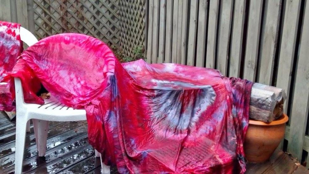 A wet length of bright, red and pale gray ice dyed fabric. The fabric is in the form of a fitted sheet, and it is draped across some backyard furniture, and it is wet, having clearly been recently rinsed out.