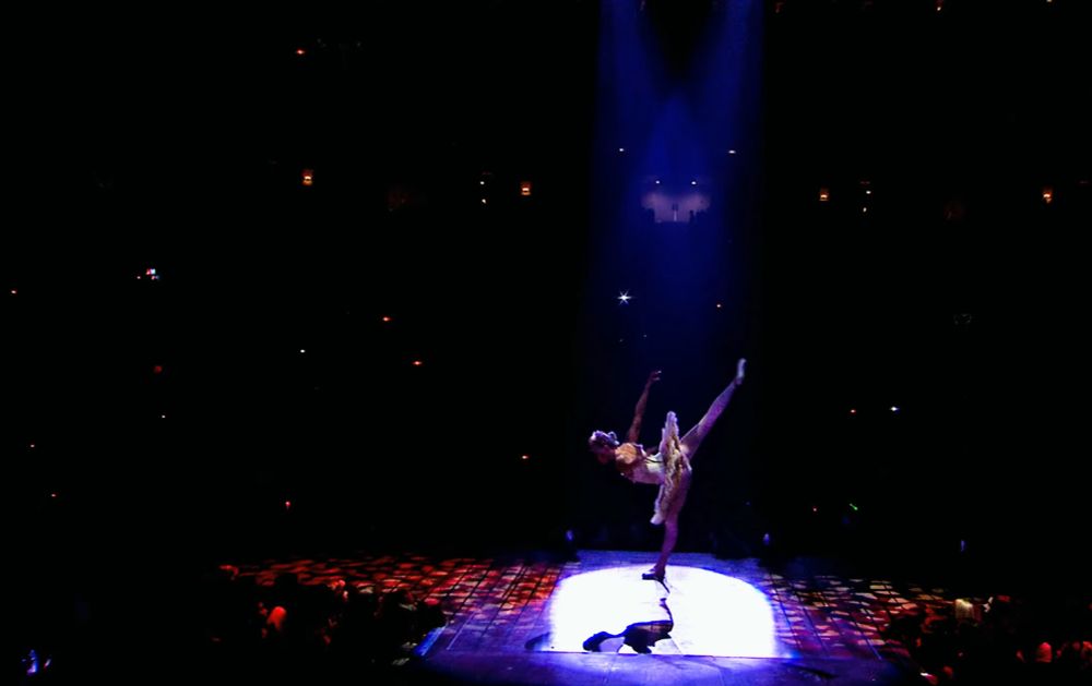 backup dancer claire callaway at the beginning of enchanted on stage at the speak now world tour. she's dancing on piano notes during the instrumental intro wearing a white dress and standing on one foot, as the other one as well as her hands are in the air. the stage spotlight is on her.