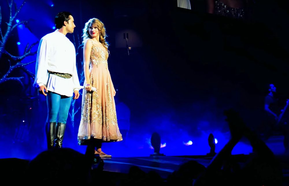 taylor swift, wearing her champagne dress, and backup dancer justin boulet, wearing a white top and blue trousers with high black boots, standing next to and looking at each other smiling moments before taylor starts singing "please don't be in love with someone else" from enchanted at the speak now world tour