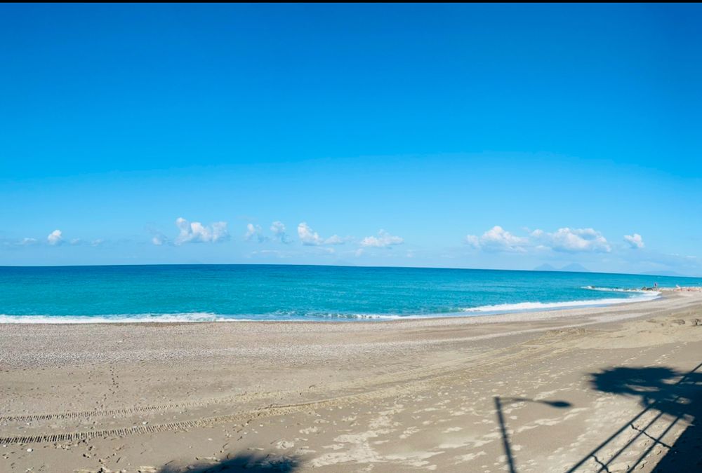 picture of the beach and sea. sea is pretty calm and water’s a few shades of blue, sky is clear with some clouds on the horizon