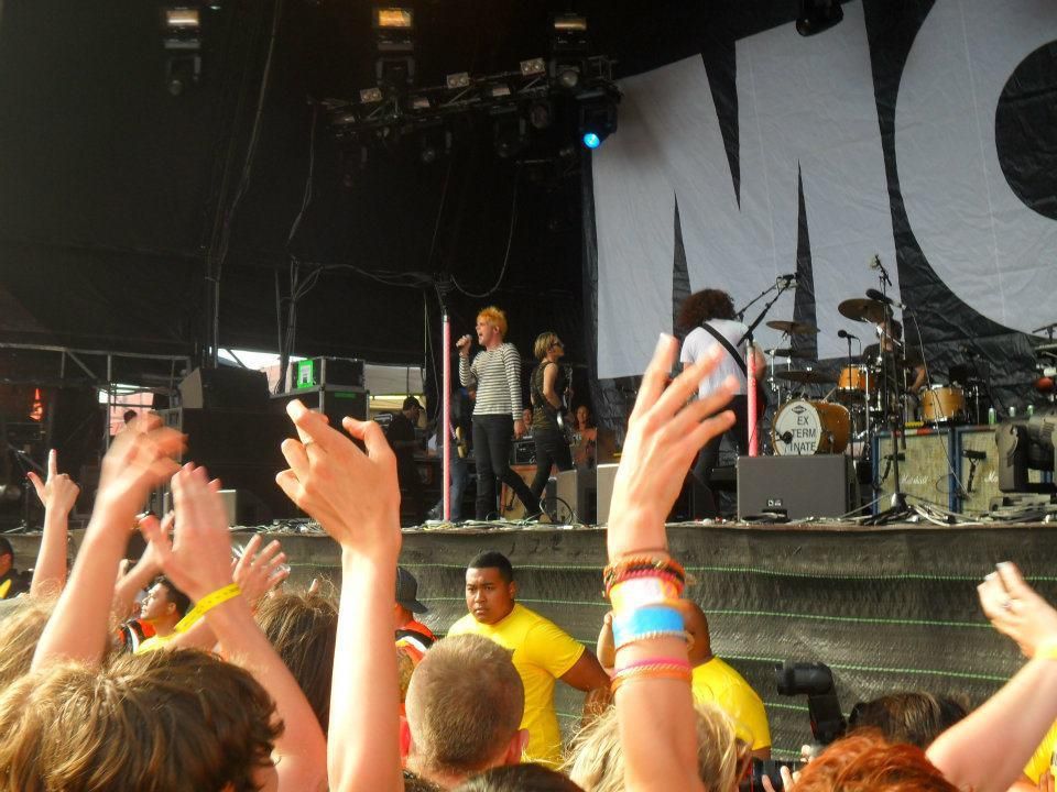My Chemical Romance performing live on an outdoor stage. Lead singer Gerard Way, with bright orange hair and a black-and-white striped shirt, holds a microphone at center stage. Other band members play guitar and drums, with a large black-and-white backdrop reading “MCR” behind them. Fans in the foreground raise their hands, while security staff in yellow shirts stand at the front of the crowd.