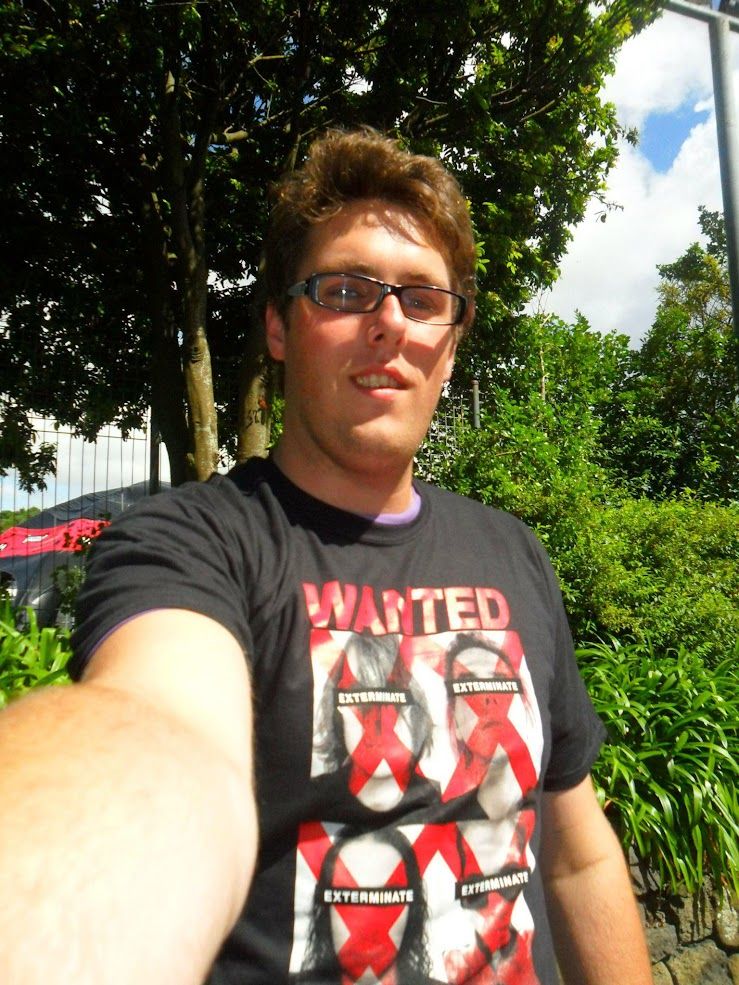 A person taking a selfie outdoors on a sunny day, wearing glasses and a black My Chemical Romance T-shirt with a “WANTED – EXTERMINATE” design featuring red X marks over faces. There are trees and greenery in the background.