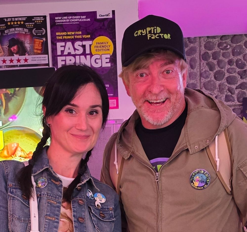 Imager of a smiling person with long dark plats and a denim jacket next to another smiling person wearing a dark beige jacket and a black cap with cryptid factor written on it.