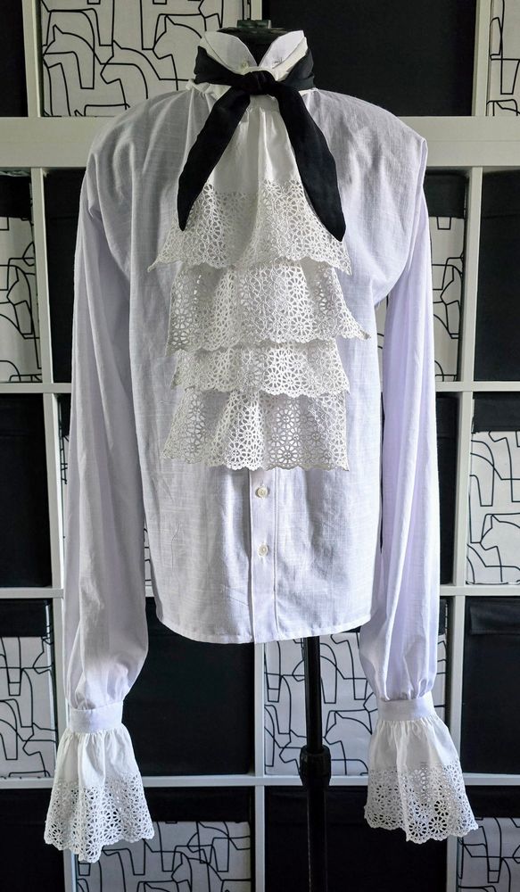 A white early 18th century style shirt on a tailors dummy with frilly broderie anglaise cuffs & matching four layer cravat. A thin black cravat is tied  over it at the throat.
