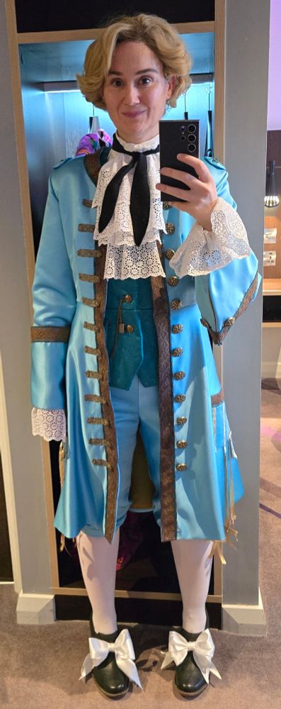 Pictur of a person dressed in Stede Bonnets outfit from the pilot episode of Our Flag Means Death.
It is a 18th century turquoise coat & breeches with bronze braiding & buttons. There is a darker turquoise waistcoat underneath.
Their legs have white stockings & black heeled shoes with white bows.
