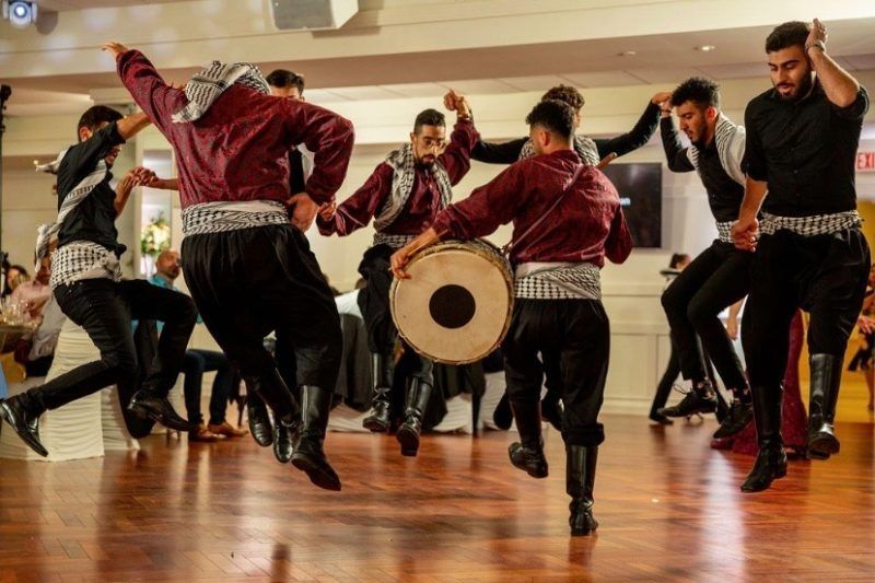Members of the Al-Asla Dabkeh group perform a traditional Levantine folk dance indoors known as Dabkeh. The dancers, wearing black and maroon outfits with Palestinian keffiyeh sashes, are captured mid-jump in a synchronized formation while one member plays a large drum. The atmosphere is festive, with onlookers seated in the background.
