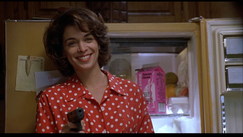 Annabella Sciorra in a red polka dot top standing in front of an open 1970s-yellow fridge, holding a gun and smiling.