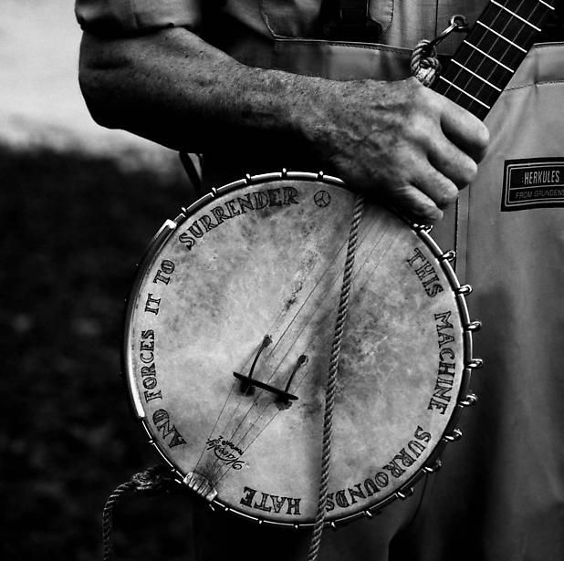 A B&W photo of a banjo. Around the circumference of the banjo face has been written THIS MACHINE SURROUNDS HATE AND FORCES IT TO SURRENDER.