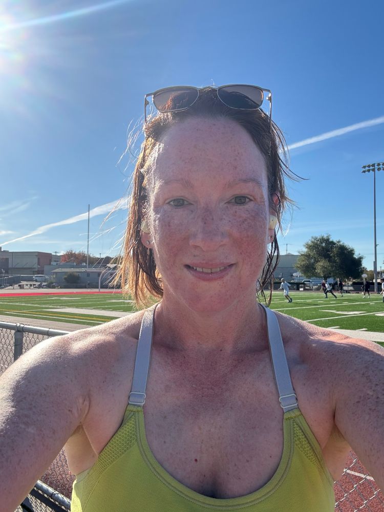 Selfie of me Sunday after 6k split run, green running top, red faced, sweaty, bright sun, dark wet hair let down from ponytail, sunglasses atop head, track and field in background, soccer game going on. 