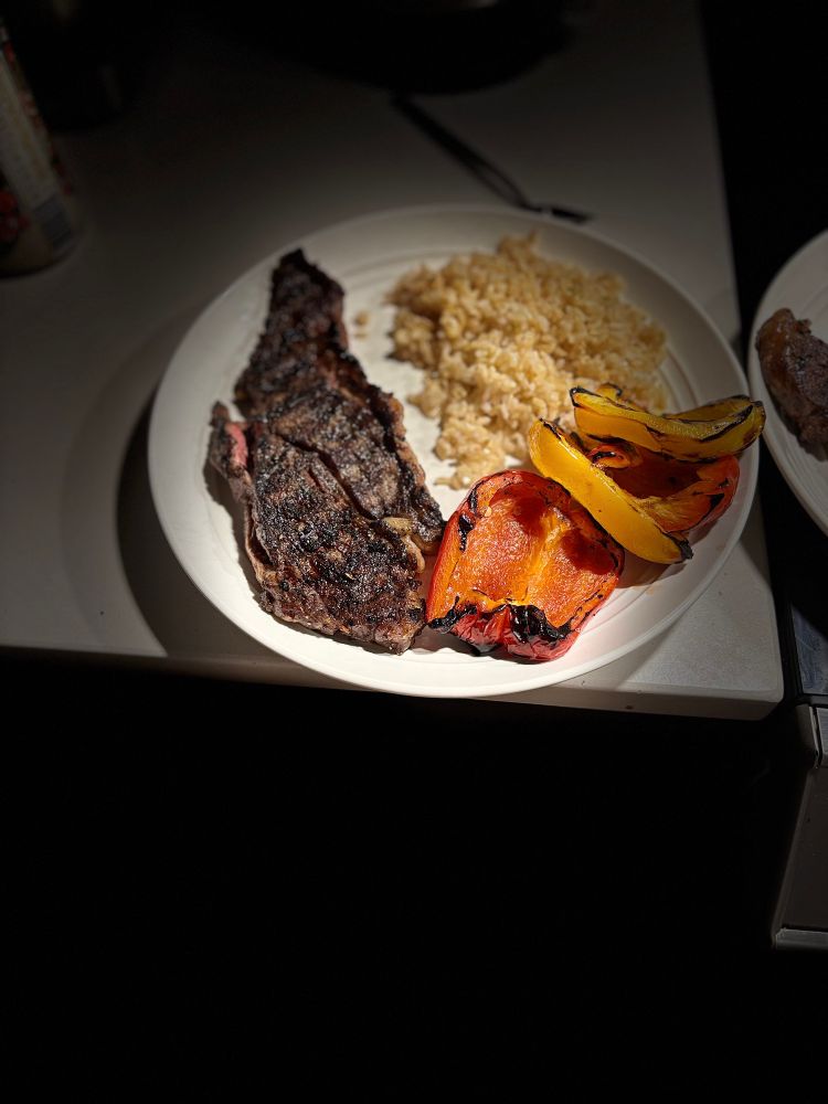 Dinner fuel. Grilled beef delmonico 🥩 (butterflied it across two portions for even grilling), grilled yellow and orange brown peppers, mound of brown rice atop white plate. This is my husband’s steak portion. 