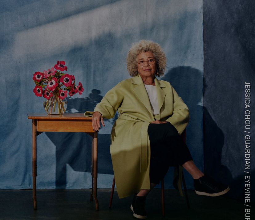 Angela Davis assise les jambes croisées fixant le photographe à côté d’une table sur laquelle repose un bouquet de fleurs dans un vase.