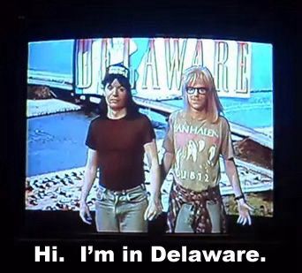 A still from the movie “Wayne’s World” showing Wayne and Garth standing awkwardly in front of a green screen with a photo of a bridge and the word Delaware. Wayne disappointingly says: “Hi, I’m in Delaware.”