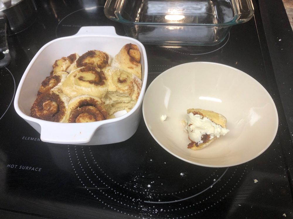 A casserole dish with 8 baked cinnamon rolls next to a plated cinnamon roll, topped with cream cheese frosting to the right. 