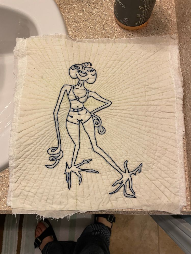 Closeup of hand embroidery and quilting - a pumpkin-headed, root-footed hottie in a bikini top embroidered in dark blue thread on a cream background. Straight lines of hand embroidery in cream thread radiate out from the hottie’s belly button. The whole piece is about 11” square.